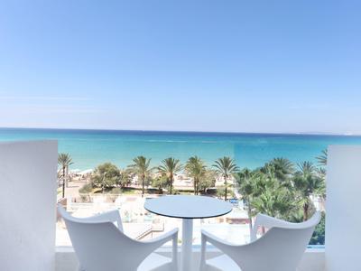 Blick von Balkon mit weißem Tisch und Stühlen auf Palmen, Strand und blaues Meer unter klarem Himmel.
