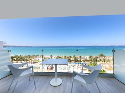 Balkon mit weißem Tisch und Stühlen, blauer Himmel, Palmen und Meer im Hintergrund.