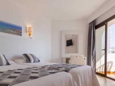 Helles Schlafzimmer mit zwei Betten, Landschaftsgemälde, Wandlampe und großem Balkonfenster.