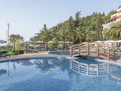 Ein Poolbereich mit einer Holzbrücke, Liegestühlen und Palmen neben einem Hotelgebäude.