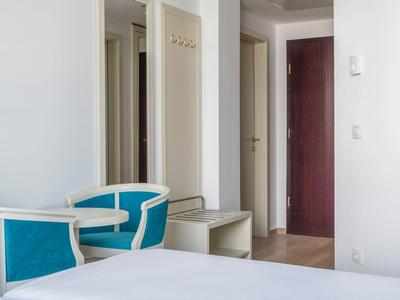 Helles Hotelzimmer mit weißem Bett, blau-weißen Stühlen und offenem Flur mit mehreren Türen.