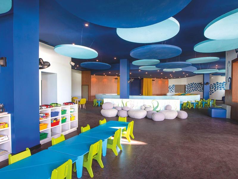 Kindgerechter Raum mit bunten Stühlen, blauen Deckenpanelen und weißen Sitzkissen.