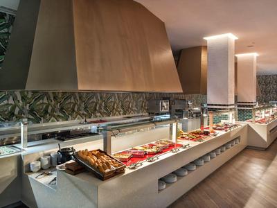 Modernes Buffet mit Metallhauben, warmen Speisen und dekorativer grüner Wand im Hintergrund.