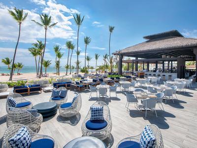Restaurant en bord de mer avec chaises blanches, coussins bleus et palmiers sous un ciel bleu.