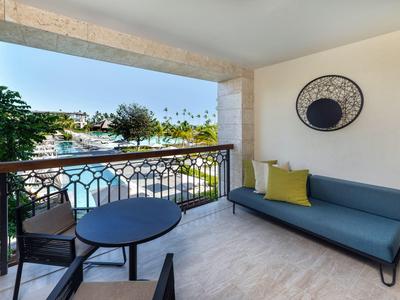 Balkon mit blauem Sofa, rundem Tisch, zwei Stühlen, hellen Wänden und Blick auf Bäume und Wasser.