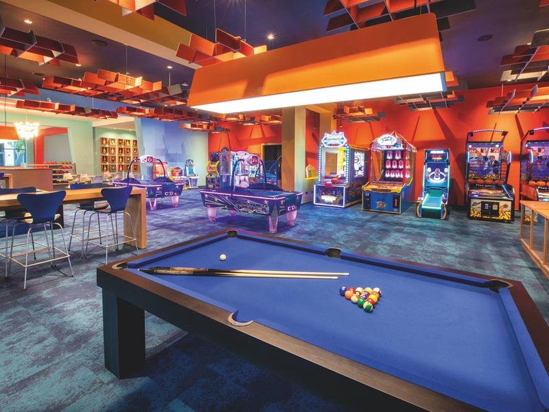 Ein Billardtisch im Vordergrund, dahinter Spielautomaten und bunte Lichter in einer Spielhalle.