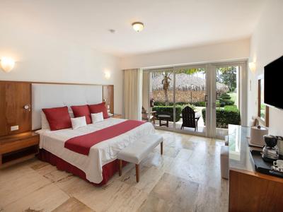 Helles Hotelzimmer mit Doppelbett, roten Akzenten, Holzmöbeln und Terrasse mit Gartenausblick.