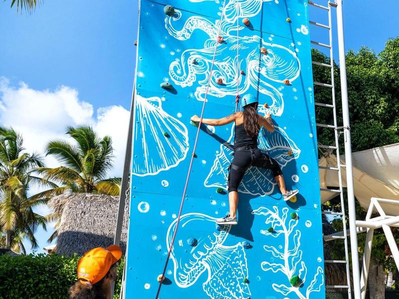 Mann klettert an blauem Kletterwand mit Meeresmotiven unter blauem Himmel und Palmen.