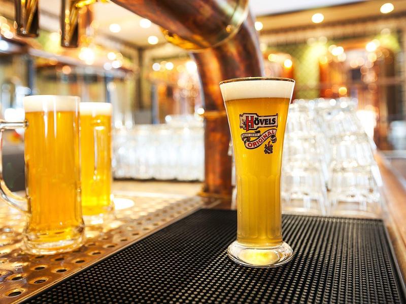 Helles Bier mit Schaum in einem Glas auf der Bar, im Hintergrund zwei Bierkrüge und Gläser.
