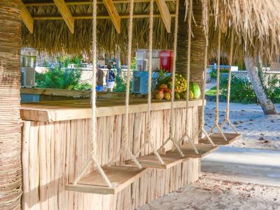 Bar mit Strohdach und vier Holzschaukeln am Strand, Sand und Palmen im Hintergrund.