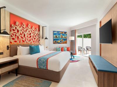 Modernes Hotelzimmer mit großem Bett, rotem Wanddekor, TV und Zugang zu Balkon mit Blick ins Grüne.