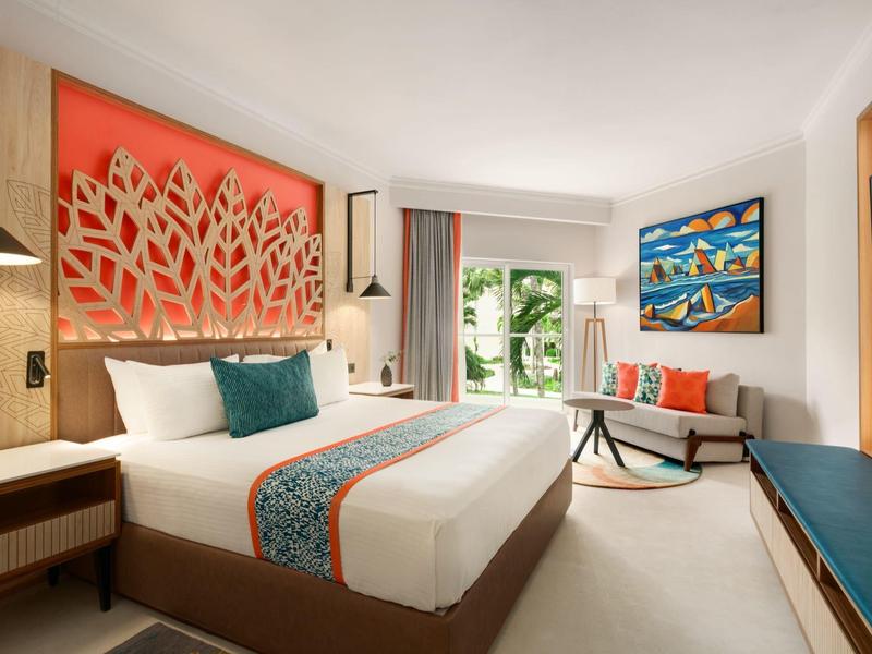 Helles Hotelzimmer mit großem Bett, buntem Wandbild, Sofa, Balkon und moderner Dekoration.