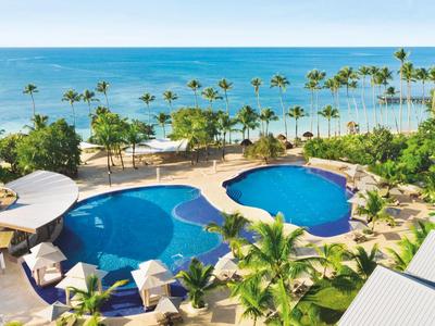 Ein großer blauer Pool mit Palmen umgeben, im Hintergrund das ruhige Meer und blauer Himmel.