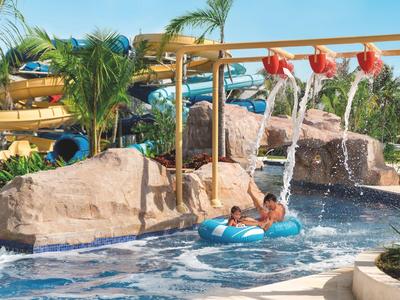 Kinder rutschen mit Reifen in blauem Wasser, umgeben von Palmen und Felsen im Freizeitpark.
