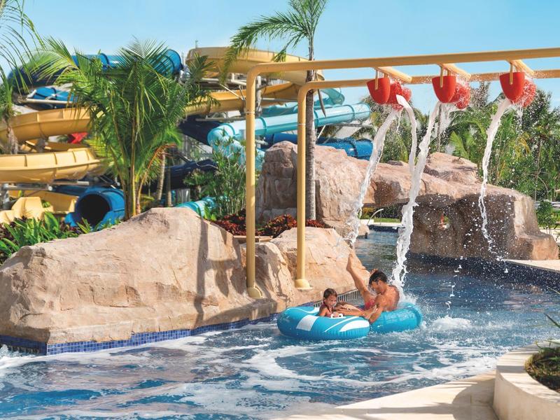 Kinder rutschen mit Reifen in blauem Wasser, umgeben von Palmen und Felsen im Freizeitpark.