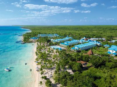 Langer Sandstrand mit türkisblauem Wasser und Hotels mit blauen Dächern in grüner Umgebung.