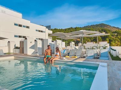 Two people sit by the edge of a pool next to a white hotel building with hills in the background.