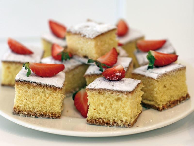 Quadratische kleine Kuchenstücke mit Puderzucker und frischer Erdbeere auf weißem Teller.