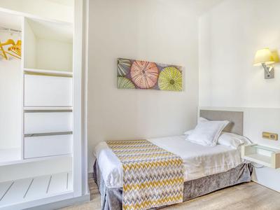 Helles, schlichtes Einbettzimmer mit Wandlampe und offener Garderobe.