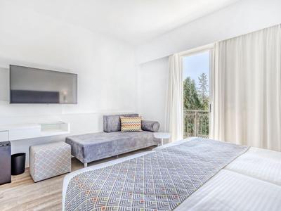 Helles Hotelzimmer mit großem Bett, Sofa, Fernseher und Balkon mit Blick ins Grüne.