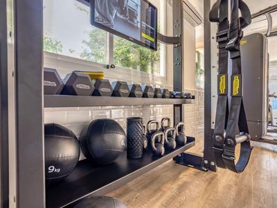 Fitness area with medicine balls, kettlebells, dumbbells, and a TRX suspension trainer in a bright room.
