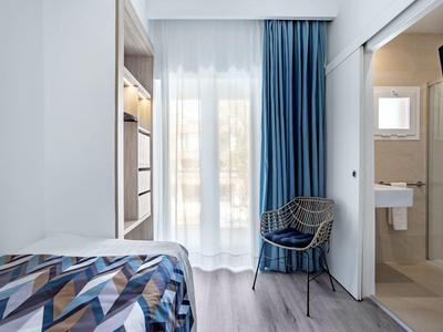 Habitación de hotel luminosa con cama, sillón junto a cortinas azules y baño abierto con lavabo.