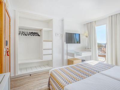 Lichte hotelkamer met twee eenpersoonsbedden, open kast en balkon met zeezicht.