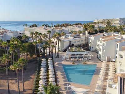 Vue aérienne d'un hôtel avec piscine, palmiers et vue sur la mer en arrière-plan.