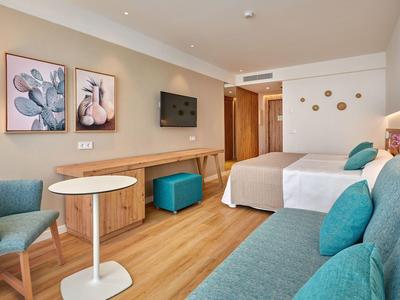 Chambre d'hôtel lumineuse avec deux lits, canapé bleu, fauteuil et décorations modernes.