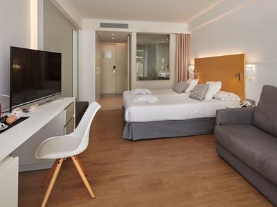 Habitación de hotel luminosa con cama doble, sofá, escritorio, televisor y suelo de madera.