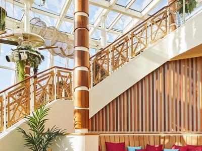 Helle Lounge mit Holztreppe, Sitzbank mit bunten Kissen und Hängepflanzen unter Glasdach.