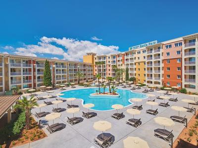 Gran piscina exterior rodeada de tumbonas y edificios de varios pisos de colores bajo un cielo azul.