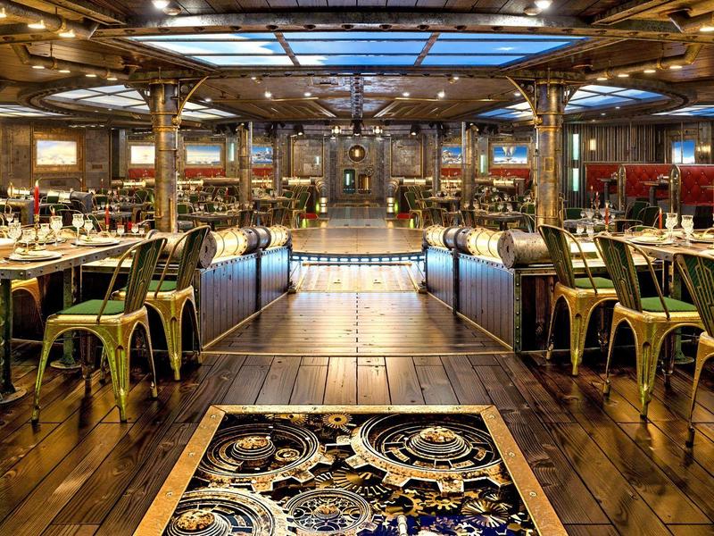 Großes, elegantes Restaurant mit Holzboden, goldfarbenen Stühlen und einem hellen Glasdach.