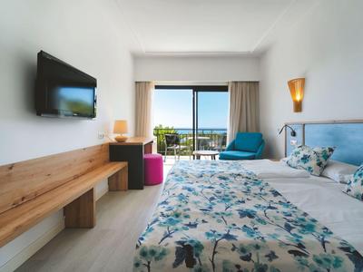 Helles Hotelzimmer mit Doppelbett, Fernseher, Sitzbereich und Balkon mit Meerblick.