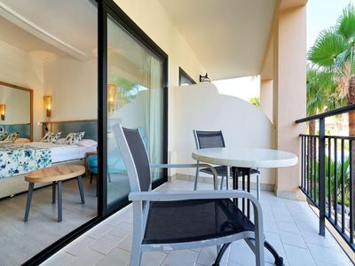 Balkon s dvěma židlemi a stolem, výhled do hotelového pokoje s manželskou postelí.