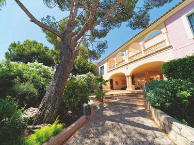 Pad leidt naar een mediterraan hotel met roze muren en grote bomen op een zonnige dag.