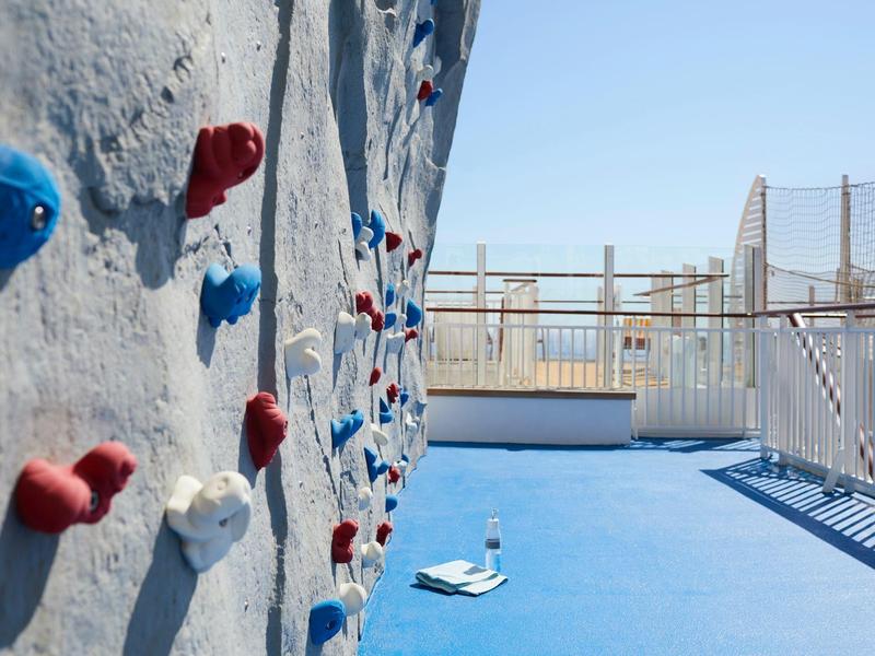 Mur d'escalade extérieur avec prises rouges et bleues sur sol bleu clair sous ciel dégagé.