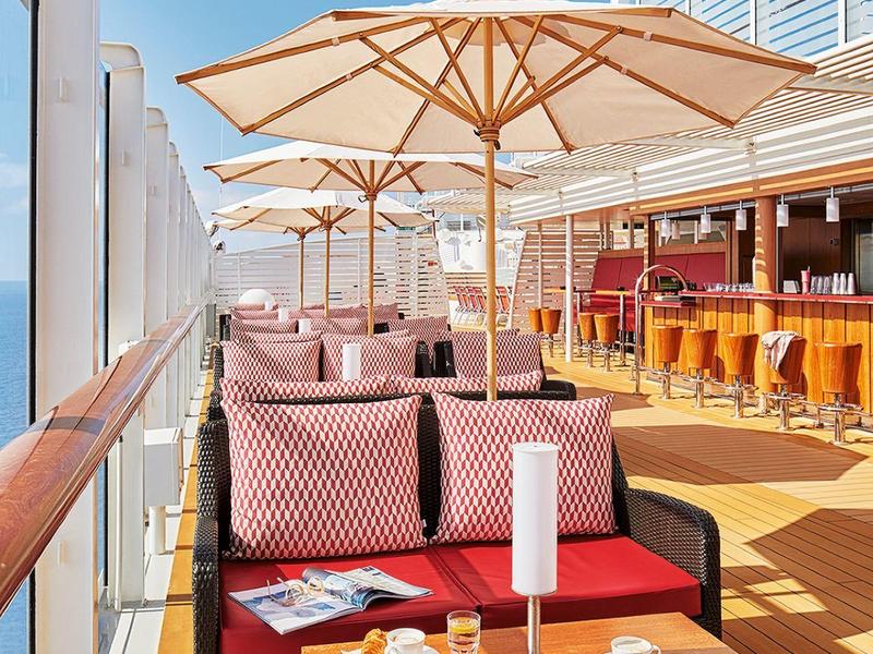 Terrasse d'hôtel avec parasols, fauteuils rouges et bar sous un ciel bleu.