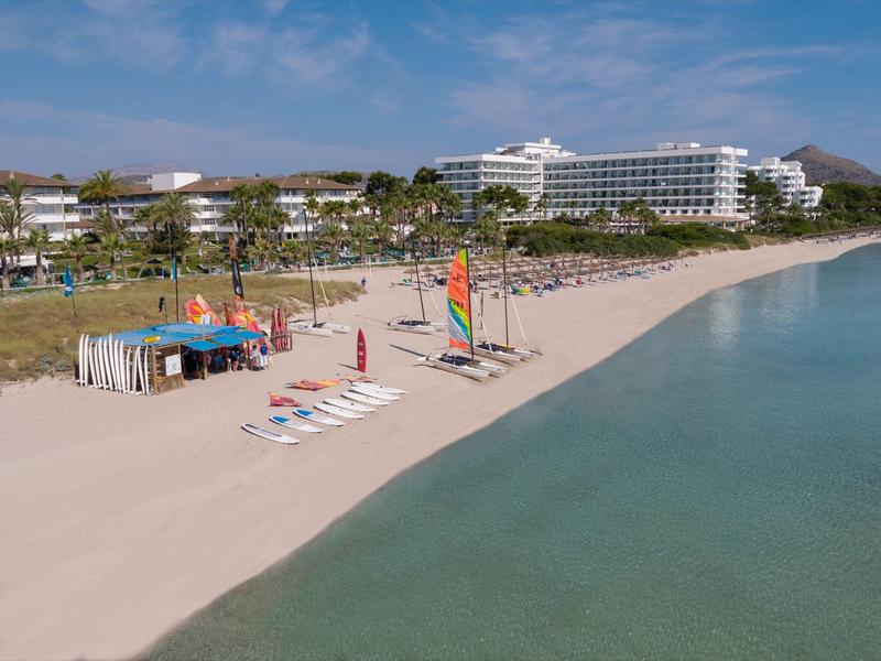 Large plage de sable avec transats, petits voiliers et tente de scène près des bâtiments d'hôtel sous un ciel bleu.