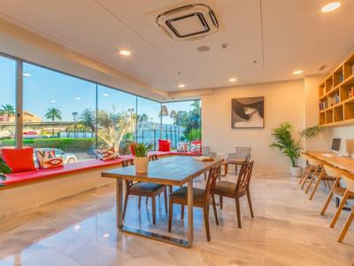 Helles Café mit Holztischen, roten Sitzpolstern, Pflanzen und großen Fenstern zum Außenbereich.