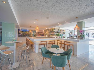 Moderner, heller Hotelbarbereich mit weißen Theken, Holzstühlen und grünen Sesseln.