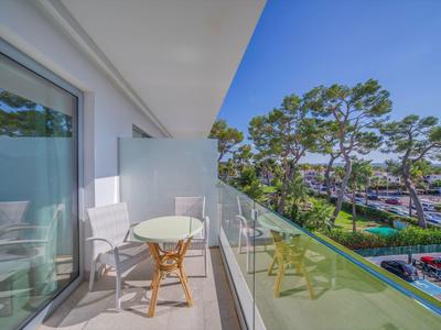 Balkon mit Glasgeländer, runder Tisch und zwei Stühlen mit Blick auf grüne Bäume.