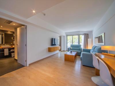 Helles Hotelzimmer mit Wohnzimmer, Sofa, TV und angrenzendem Bad mit Waschbecken.