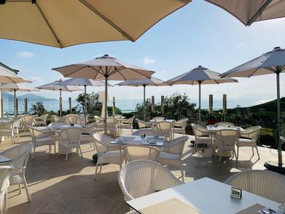 Terrasse mit weißen Tischen und Stühlen, vielen Sonnenschirmen und Blick auf das Meer.