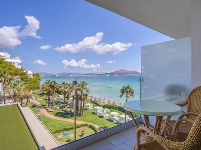 Blick von einem Balkon mit Tisch und Stühlen auf tropischen Garten und Meer mit Bergen im Hintergrund.