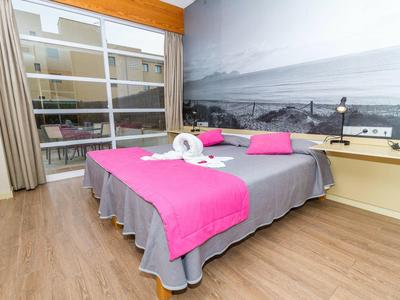 Modern hotel room with double bed, gray bedding and pink accents, window overlooking terrace.