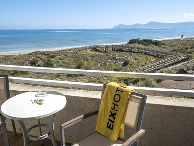 Balcony with table and chair, sea view and sandy dunes in the background under clear sky.