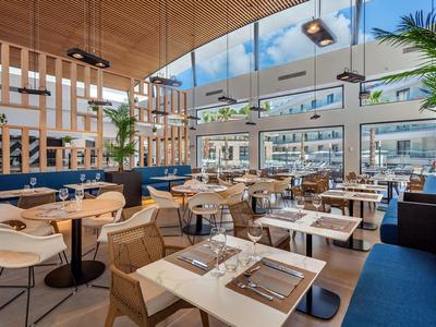 Espace restaurant d'hôtel moderne avec grandes fenêtres, banquettes bleues et plantes.