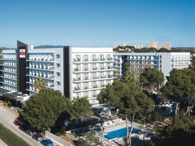 Modern hotel surrounded by trees with a pool and balconies under a clear blue sky.