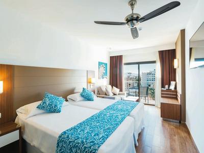 Bright hotel room with two beds, blue accents, ceiling fan, and balcony view.